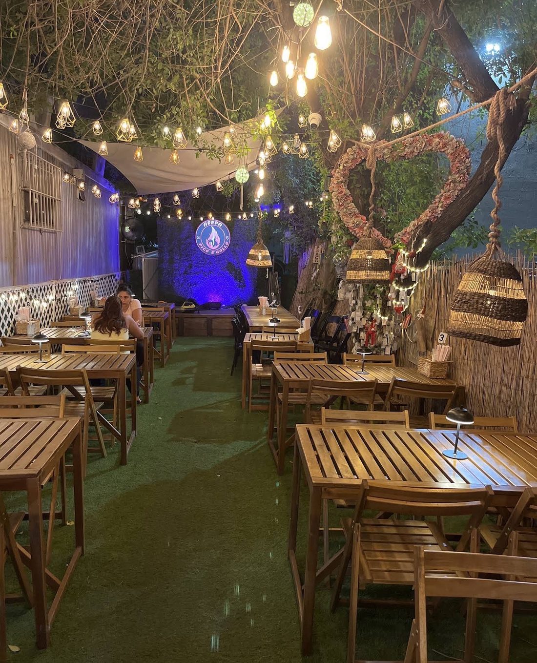 The Pico e Gallo BBQ dining patio at night with string lights, a heart decoration, the wishing tree and the “Arepa Pico e Gallo” neon sign